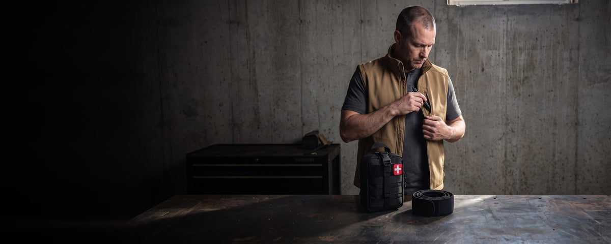 A man in a tan zip vest over a dark tee, one hand at the chest zipper, beside a first aid pouch and a rolled black belt on a steel workbench
