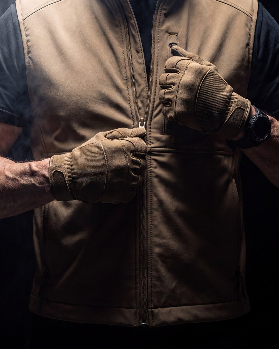 Man in tan gloves zipping a beige softshell carry vest over a dark tee, dark studio