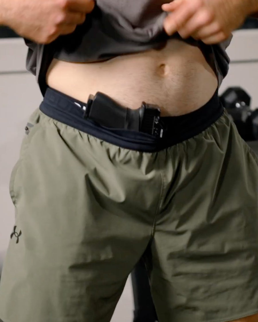 Man lifting a gray tee to show a pistol in a waistband holster over a black waistband, olive gym shorts, dumbbells behind