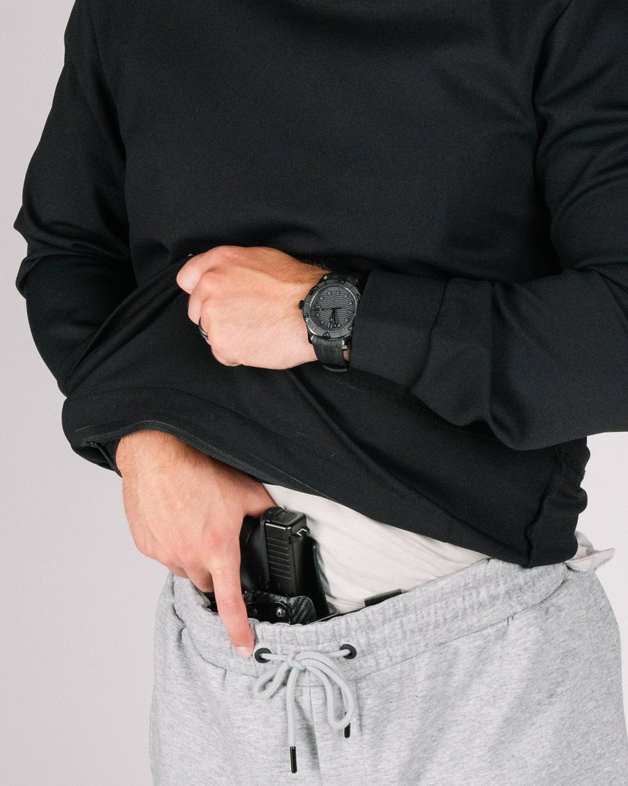 Man lifting the hem of a black hoodie to show a pistol in a waistband holster over gray sweatpants