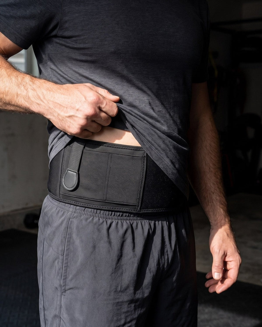 Man lifts a charcoal t-shirt hem to show a black belly band with a holster pocket and strap, above grey shorts.